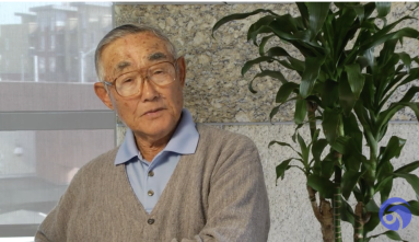 Jimmy Ko Fukuhara wearing a blue collared shirt and a tan sweater sits next to a plant and speaks of his experience in the sugar beet farms
